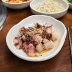 Flache weiße Schale mit klein geschnittenen Tintenfischtentakeln, Sesam und Metallstäbchen