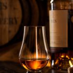 Tulpenförmiges Nosingglas mit bernsteinfarbenem Whisky auf dunklem Holztresen, dahinter unscharf ein Fass