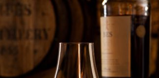 Wie schmeckt Whisky? Tulpenförmiges Nosingglas mit bernsteinfarbenem Whisky auf dunklem Holztresen, dahinter unscharf ein Fass