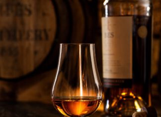 Wie schmeckt Whisky? Tulpenförmiges Nosingglas mit bernsteinfarbenem Whisky auf dunklem Holztresen, dahinter unscharf ein Fass
