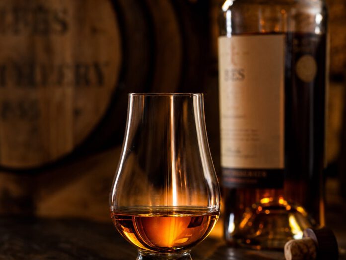 Tulpenförmiges Nosingglas mit bernsteinfarbenem Whisky auf dunklem Holztresen, dahinter unscharf ein Fass