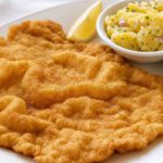 Wiener Schnitzel mit goldgelber, gewellter Panade auf weißem Teller, daneben Zitronenspalte und Kartoffelsalat