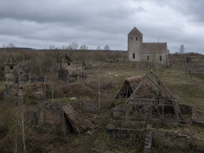 Verfallene Fachwerkhäuser und eingestürzte Dächer eines verlassenen Dorfes, verwilderte Felder und eine einsam stehende Steinkirche unter grauem Himmel