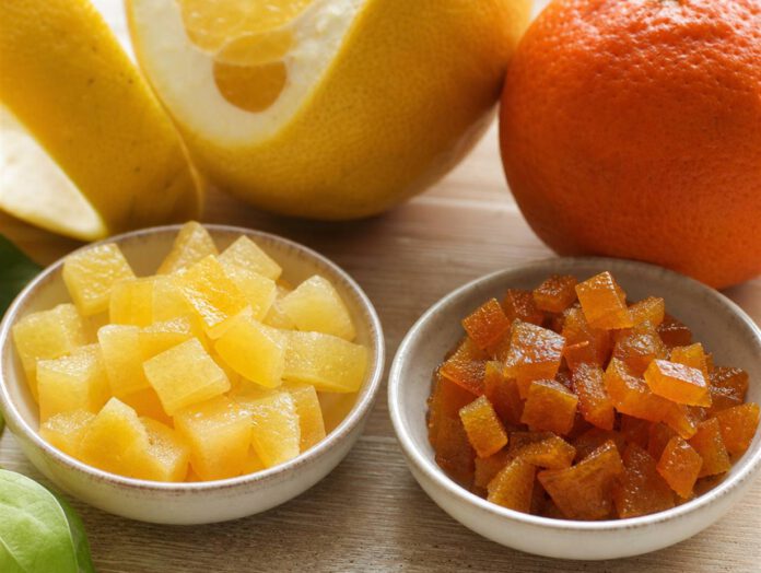 Zwei Schalen nebeneinander, links helle Würfel Zitronat, rechts bräunlich-orange Würfel Orangeat, dahinter eine dickschalige Zitrusfrucht und eine Orange