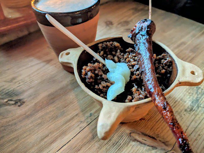 Buchweizengrütze mit Grieben und einer Haube Schmand in einer Tonschale, dazu eine gegrillte Wurst am Holzspieß