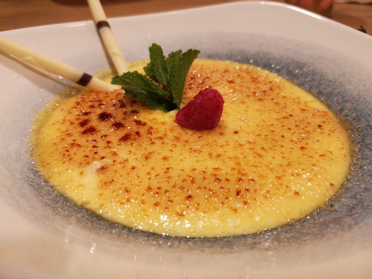 Crème brûlée mit aufgebrochener Karamellkruste, Minzblatt und Himbeere auf blauem Teller