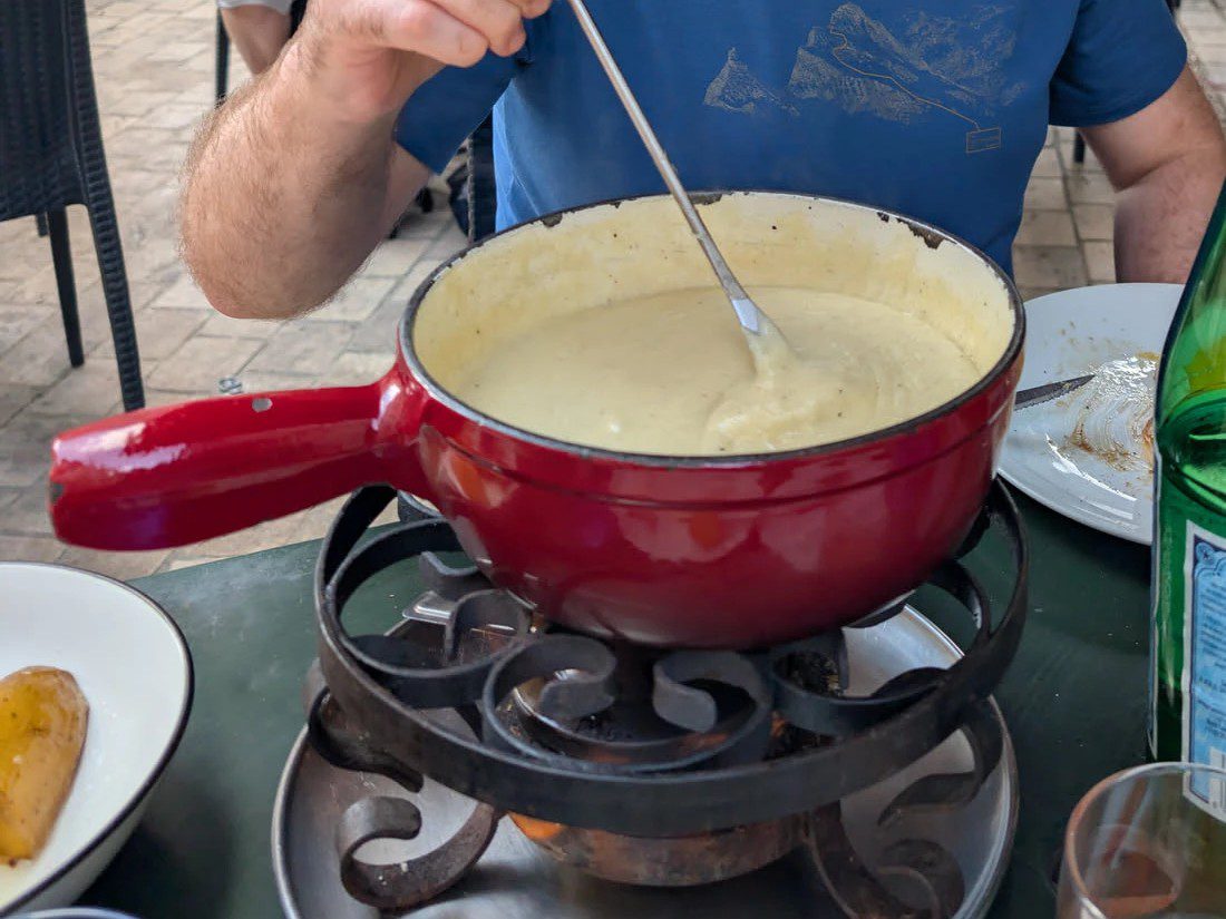 Rotes Caquelon mit geschmolzenem Käsefondue auf einem Rechaud, eine Hand führt die Fonduegabel