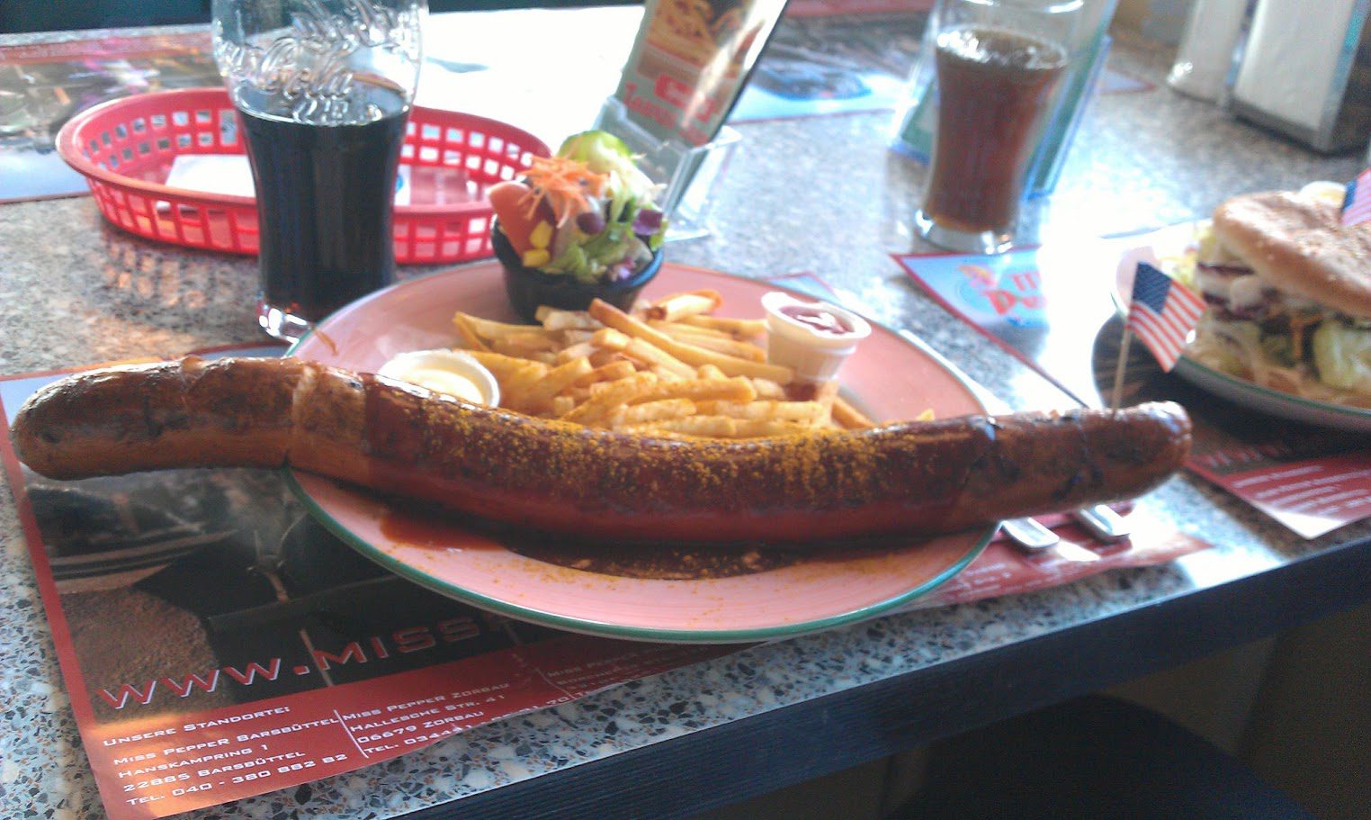Übergroße Currywurst mit Currypulver auf einem Teller, daneben Pommes frites und ein Salatschälchen