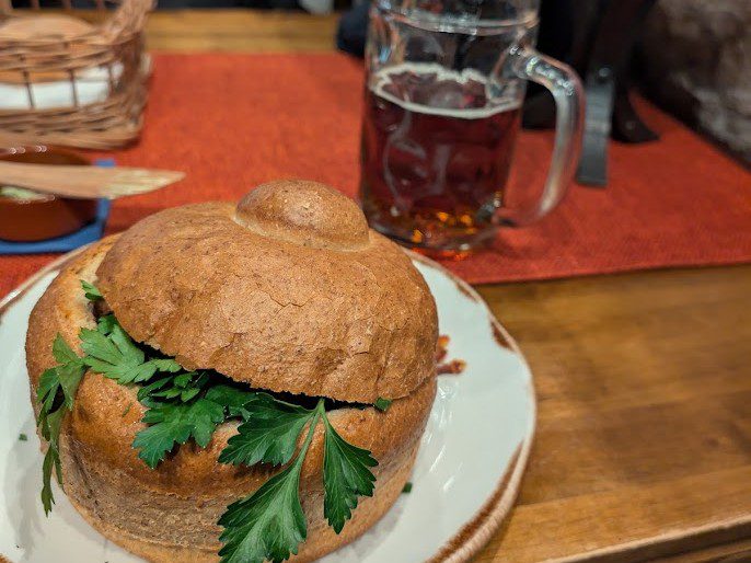 Suppe in einem ausgehöhlten runden Brotlaib mit Deckel und Petersilie, dazu ein Glas dunkles Bier