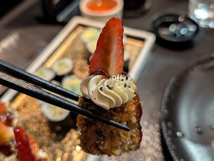 Frittierte Sushi-Rolle mit Frischkäsetupfen und Erdbeere, gehalten mit schwarzen Essstäbchen