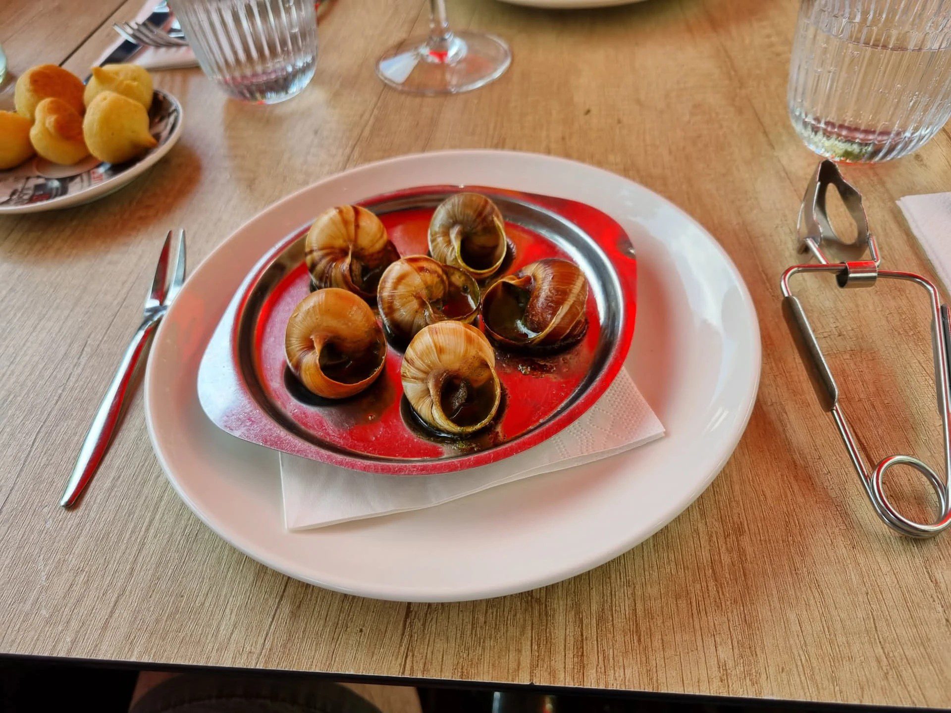 Sechs Weinbergschnecken in Kräuterbutter auf einem roten Escargot-Teller, daneben eine Schneckenzange